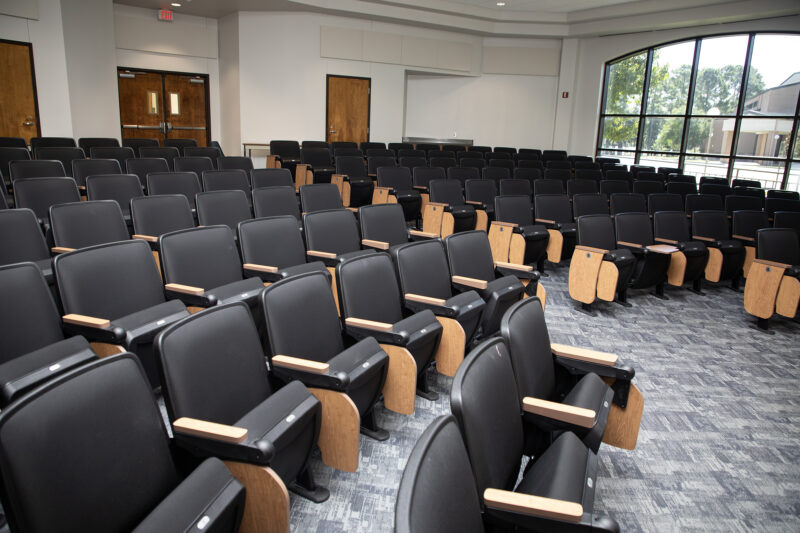 Lecture hall with empty black chairs, wooden armrests, grey carpet, large right-side windows, and sunlight streaming in.