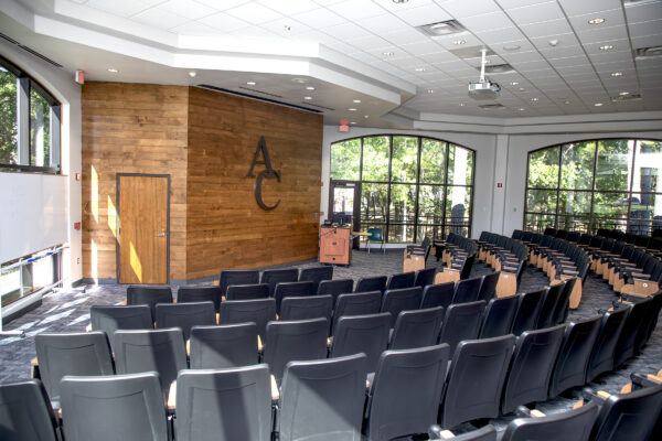 Lecture hall with black chairs facing a wooden stage labeled AC, large sunlit windows, and a ceiling-mounted projector.