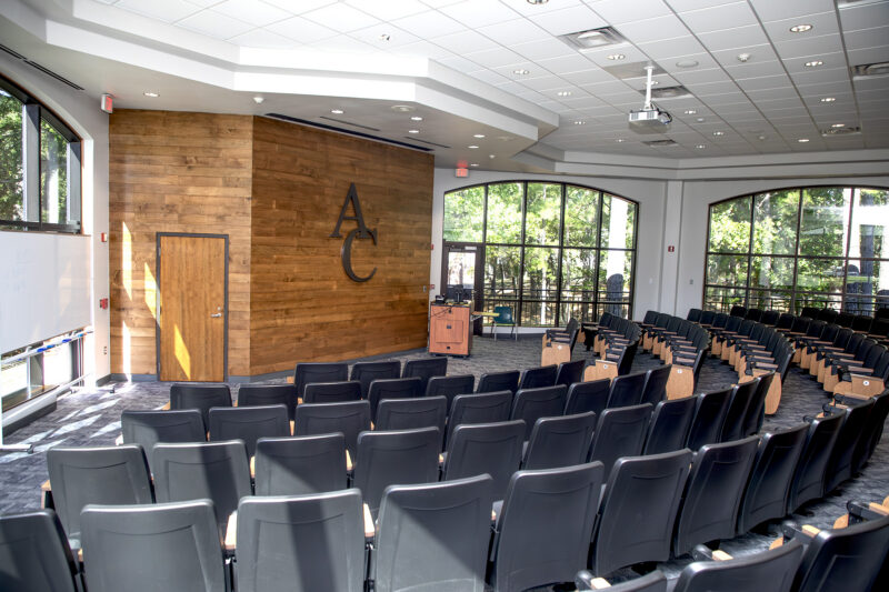 Lecture hall with black chairs facing a wooden stage labeled AC, large sunlit windows, and a ceiling-mounted projector.