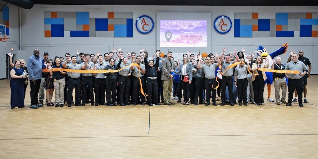 Group in gym holds orange ribbon, smiling. Some wear uniforms, others casual clothes; mascot and screen in background.