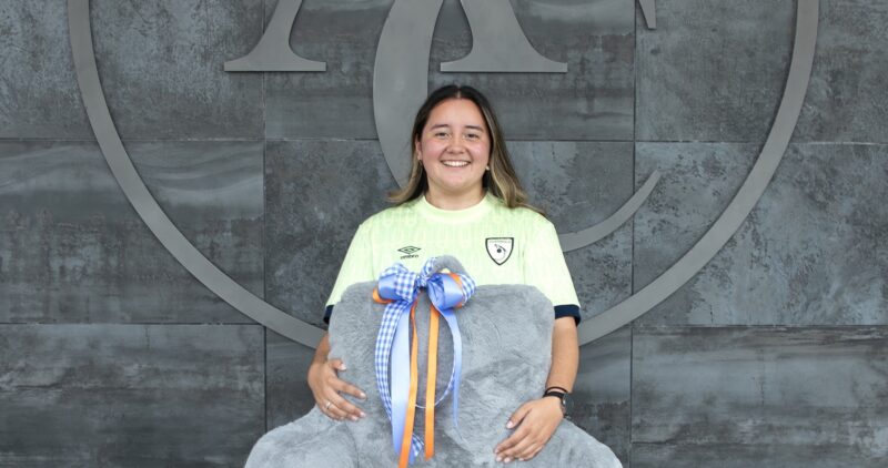 Person in a pale green sports jersey smiles, holding a gray stuffed elephant with a colorful ribbon bow; large logo on wall behind.
