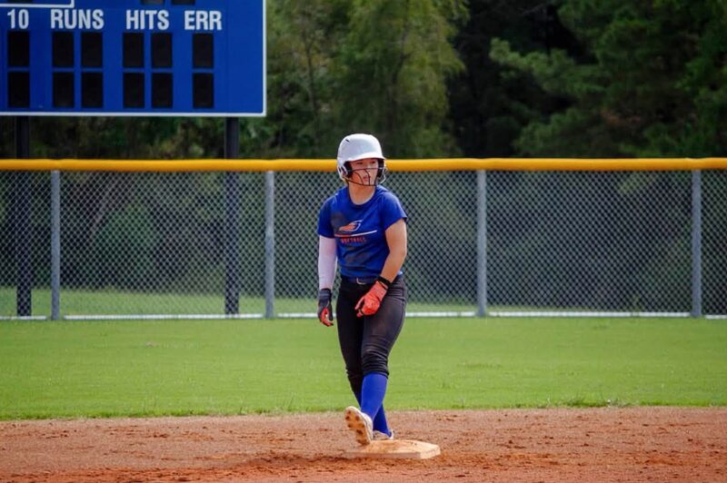Softball player in blue uniform and helmet stands on base, glancing sideways; scoreboard and trees are visible behind her.