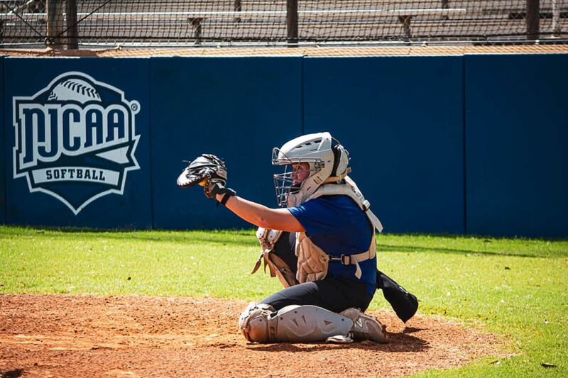 Softball catcher in full gear kneels on dirt, glove extended; blue NJCAA Softball sign on fence in background.