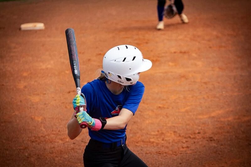 Youth baseball player in blue shirt and white helmet grips black bat, ready to hit, with another player in the background.