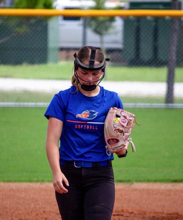 Softball player in blue shirt, black pants, and face mask walks on field with glove; background shows fence, grass, and parked cars.