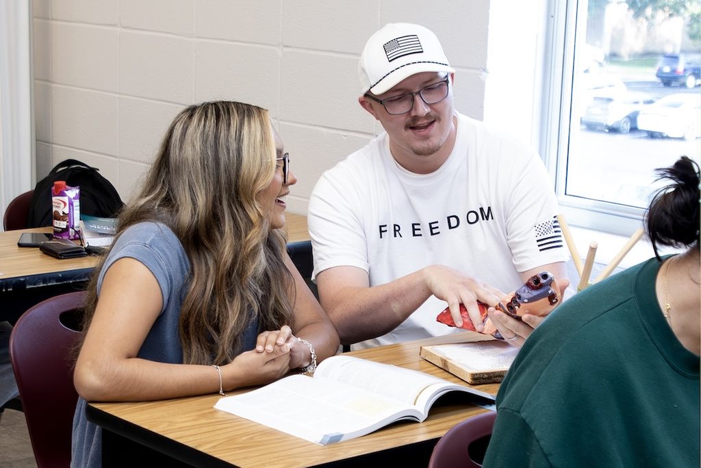 Two students smile at a phone together at a classroom desk with books, a drink, supplies, and natural light from a window.
