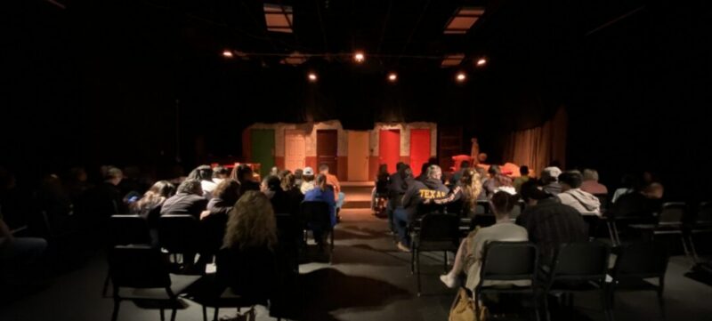 Audience in a dim theater faces a stage with four colorful doors and dramatic lighting, awaiting a performance.