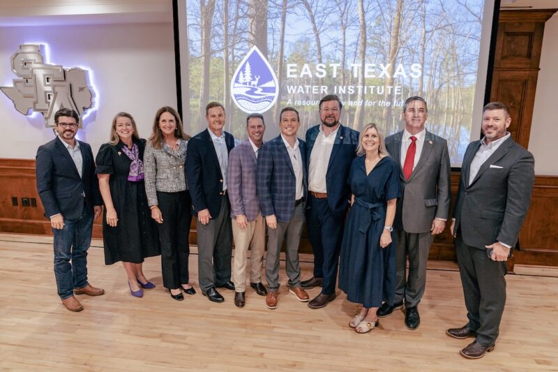 Eleven people in business attire stand side by side indoors, smiling in front of an East Texas Water Institute screen with a forest and lake.