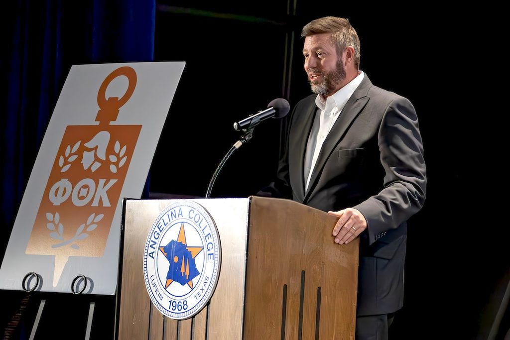 A man in a suit speaks at a podium featuring an Angelina College seal, positioned beside a Phi Theta Kappa logo sign.
