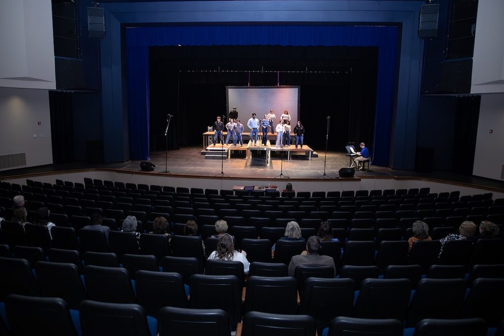 Performers standing on a theater stage with a pianist at the side, seen from the audience seating area.