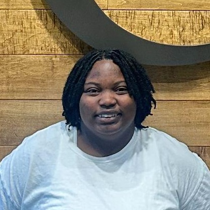 Person with short, black twisted hair smiles at the camera, wearing a white shirt in front of a wooden wall and dark circular object.