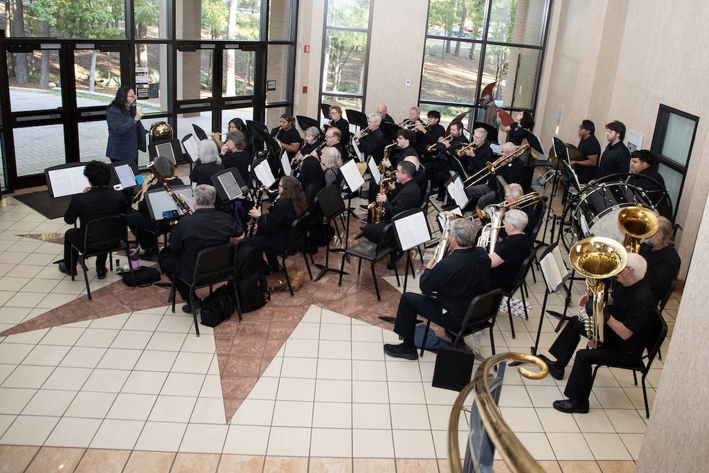 A conductor leads a concert band performing in a spacious lobby with floor-to-ceiling windows.