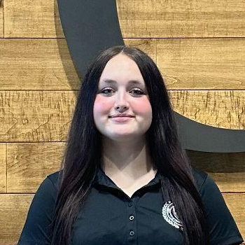 Young woman with long brown hair and a black collared shirt stands before a wooden wall featuring a curved black design.