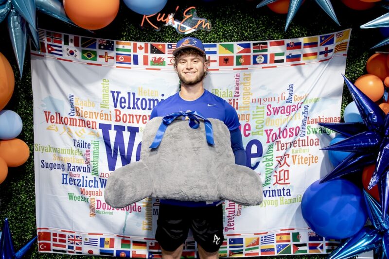 Smiling man in athletic gear holds a large stuffed whale tail before a colorful Welcome banner with world flags and balloons.