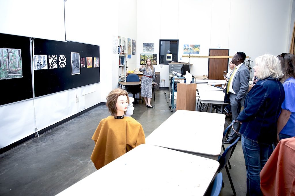 A woman speaks to a group in an art classroom, with various artworks displayed on a chalkboard nearby.