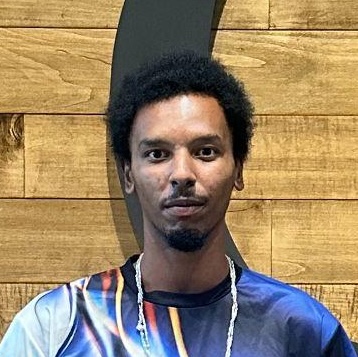 Man with short curly hair and a goatee stands before a wooden wall, wearing a colorful shirt and chain necklace.