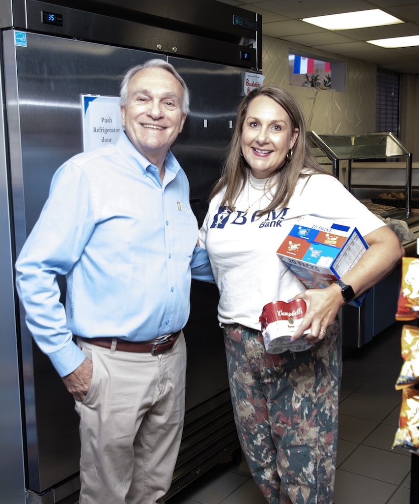 A man in a light blue shirt and a woman in a BGM Bank t-shirt hold food boxes before a commercial refrigerator.