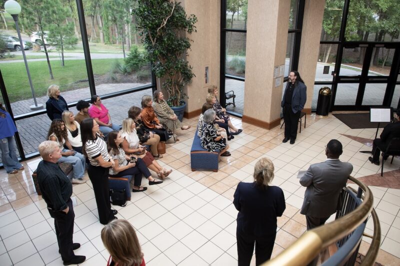 A man in a blazer addresses a seated audience in a bright, modern lobby with large windows.