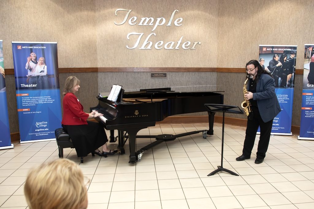 A woman in a red blazer plays a grand piano accompanied by a man on saxophone in front of a Temple Theater sign.