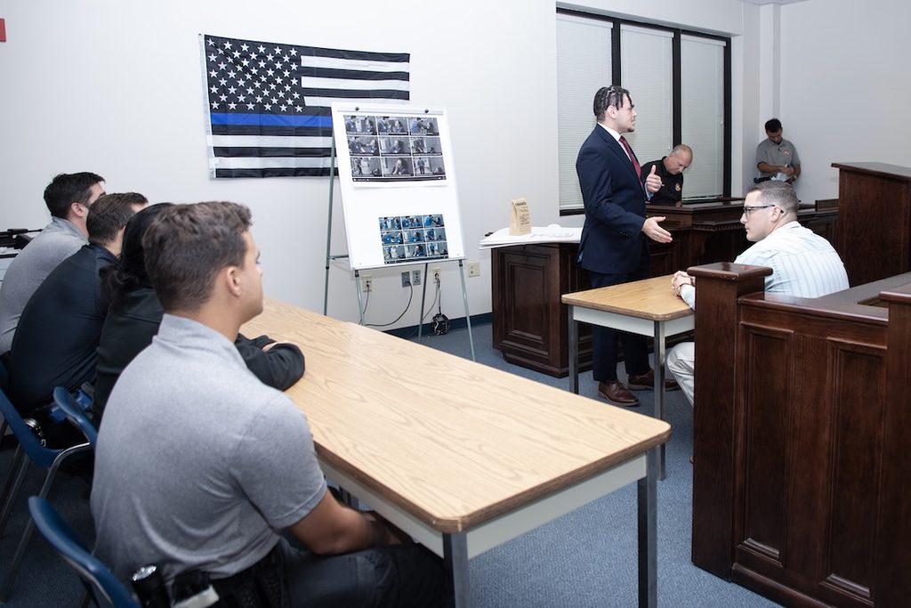 A man in a suit addresses a group in a room featuring a thin blue line flag and a display board of photographs.