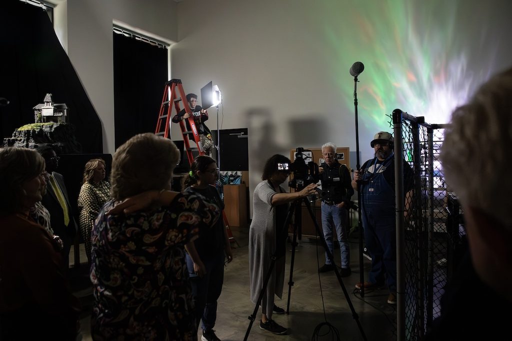 A film crew records a miniature house model in a studio under specialized lighting.