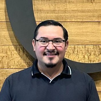 Man with glasses and short dark hair in a black polo shirt smiles in front of a wooden wall with a dark circular design.
