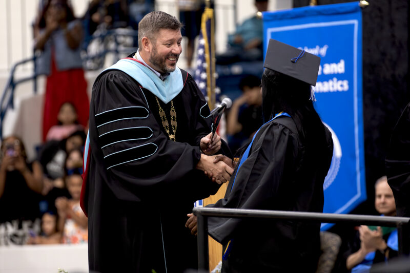 A university official in academic regalia shakes hands with a graduate on stage during a commencement ceremony.