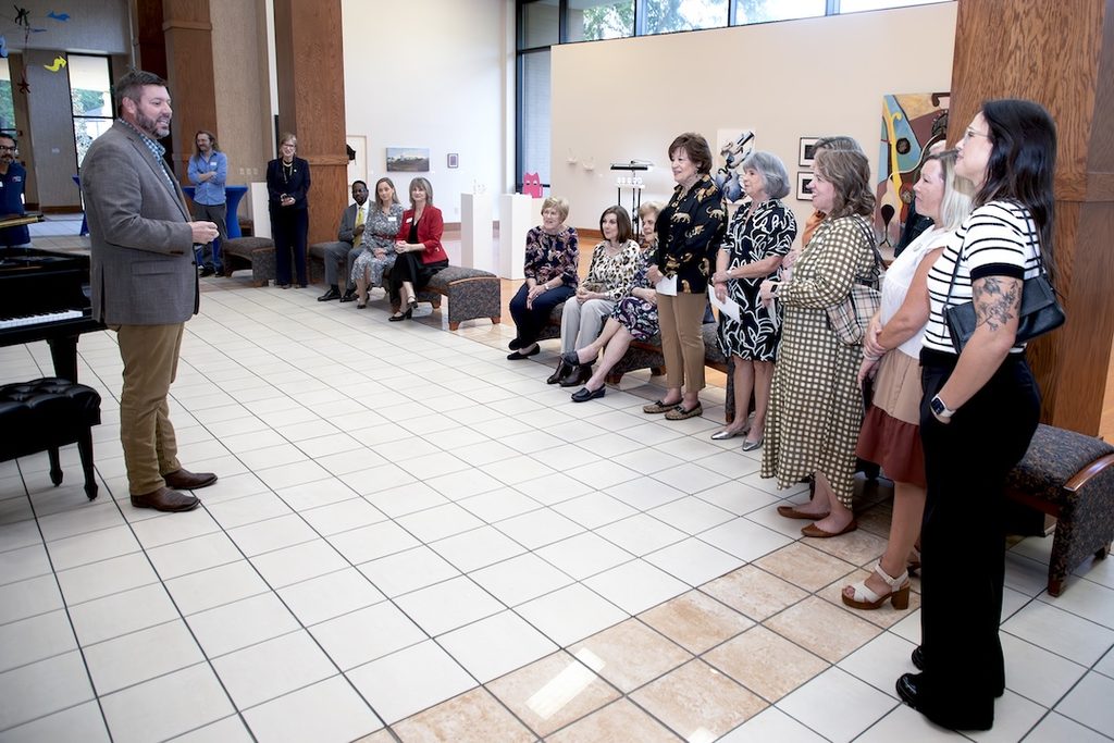 A man in a suit jacket speaks to a group of people gathered in an art gallery.