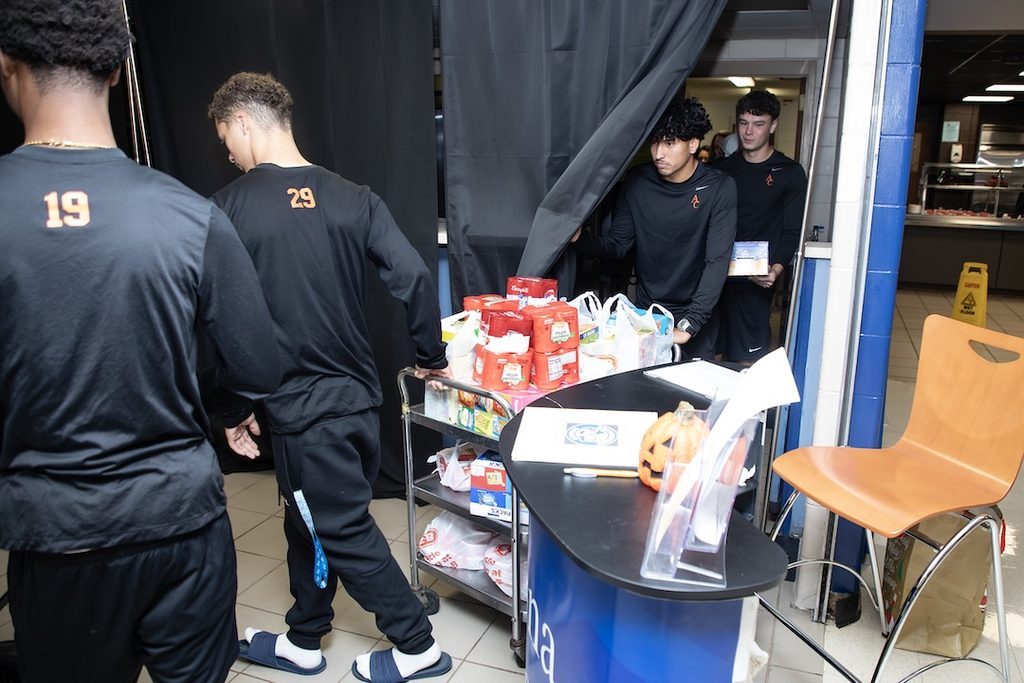 Four young men in matching black athletic long-sleeve shirts push a cart filled with groceries and a small pumpkin.
