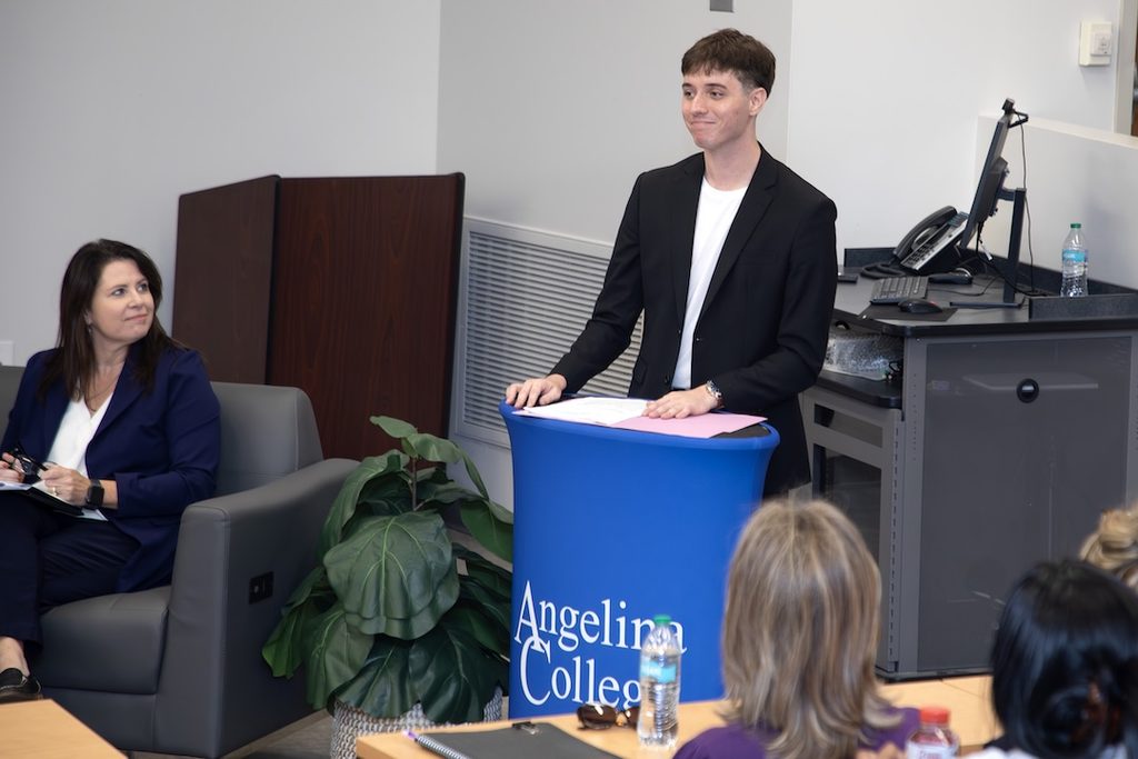 A young man in a black blazer speaks from behind a blue Angelina College lectern to a seated audience.