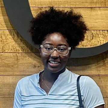 Person with curly hair, clear glasses, and a light blue top smiles before a wooden wall featuring a large dark circle.